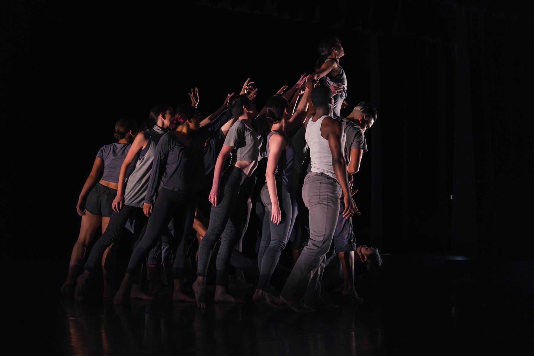 Dance students under a bright spotlight in a dark stadium pose tightly together on stage.